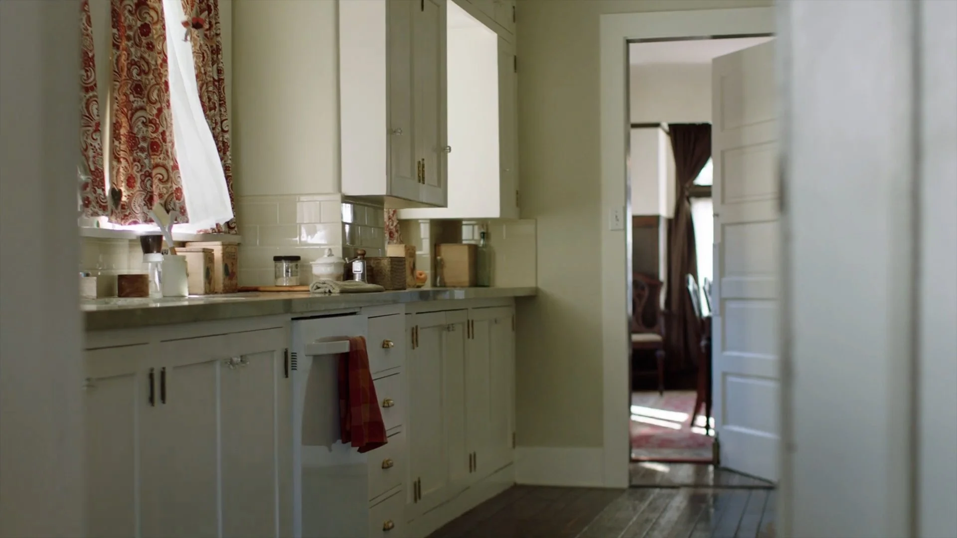 A vintage kitchen with white cabinets, a window with patterned red and white curtains, and a view through an open door into another room with dark curtains and chairs.