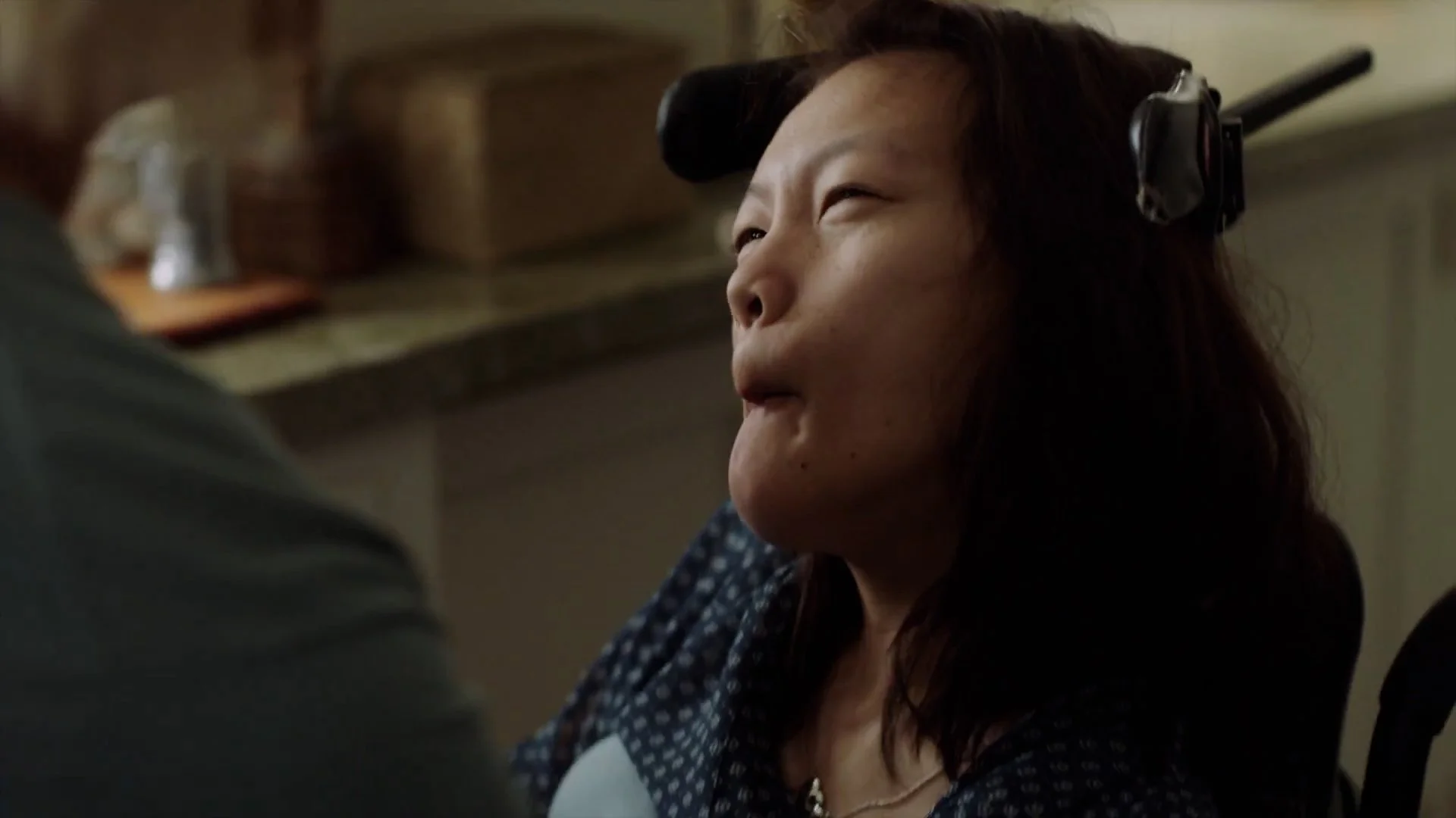 A woman with a hair roller on her head sitting in a kitchen, reacting emotionally, possibly crying or on the verge of tears.