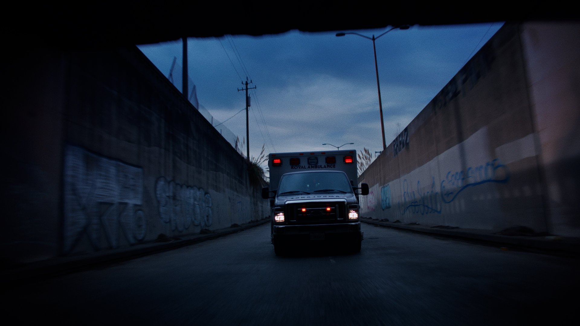A captured view of an ambulance driving through a narrow alley at dusk, with graffiti-covered concrete walls on either side and utility poles visible above.