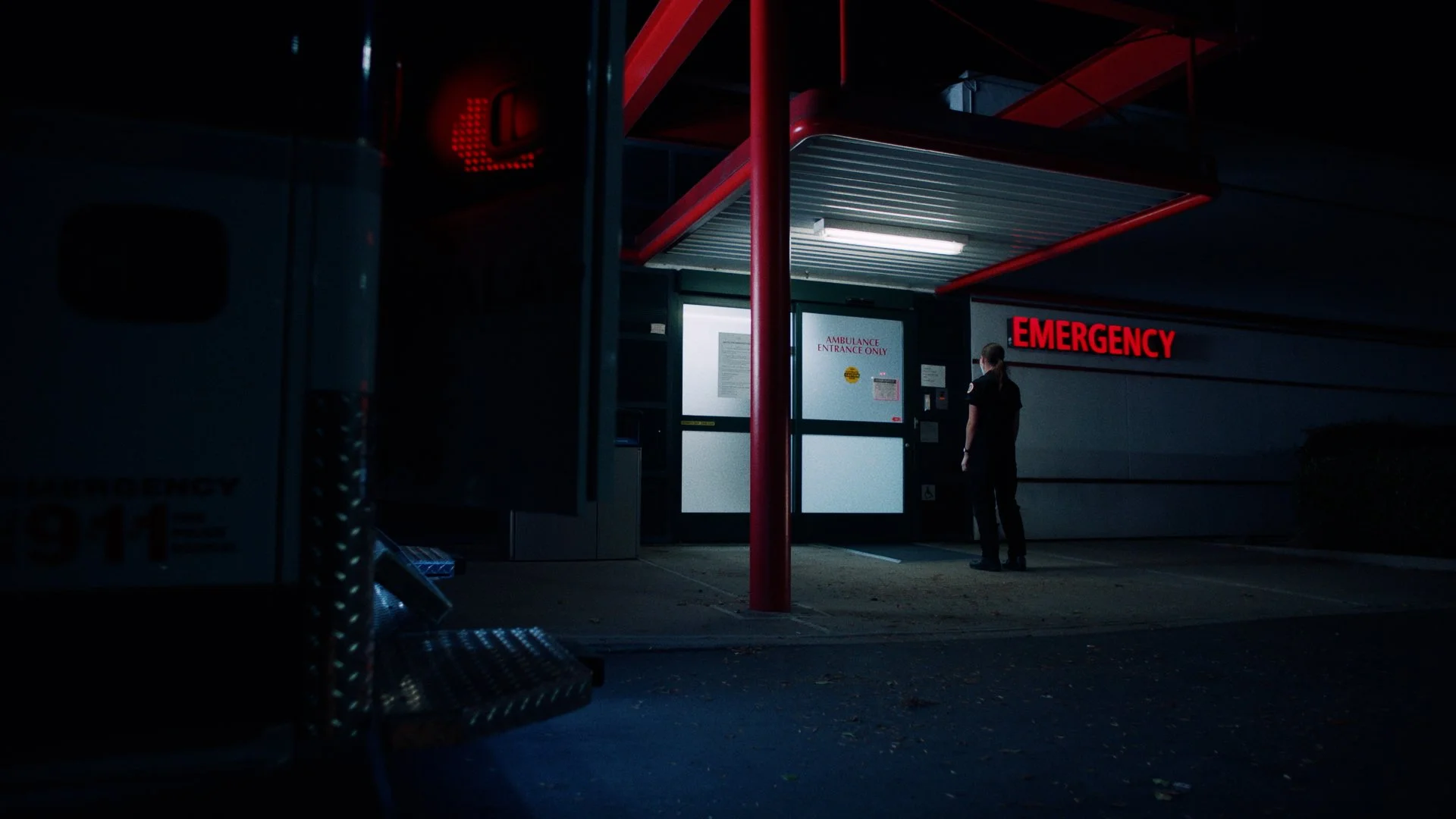 A person standing outside the emergency entrance of a hospital at night, illuminated by red and white lights, with an ambulance partially visible on the left.