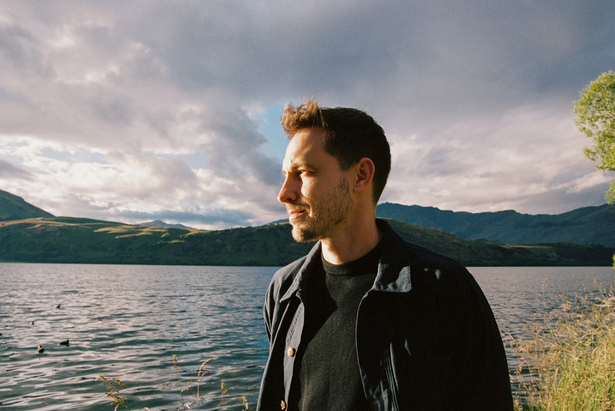 A man with short dark hair and a beard standing by a lake, looking to the left, with mountains and cloudy sky in the background during daylight.