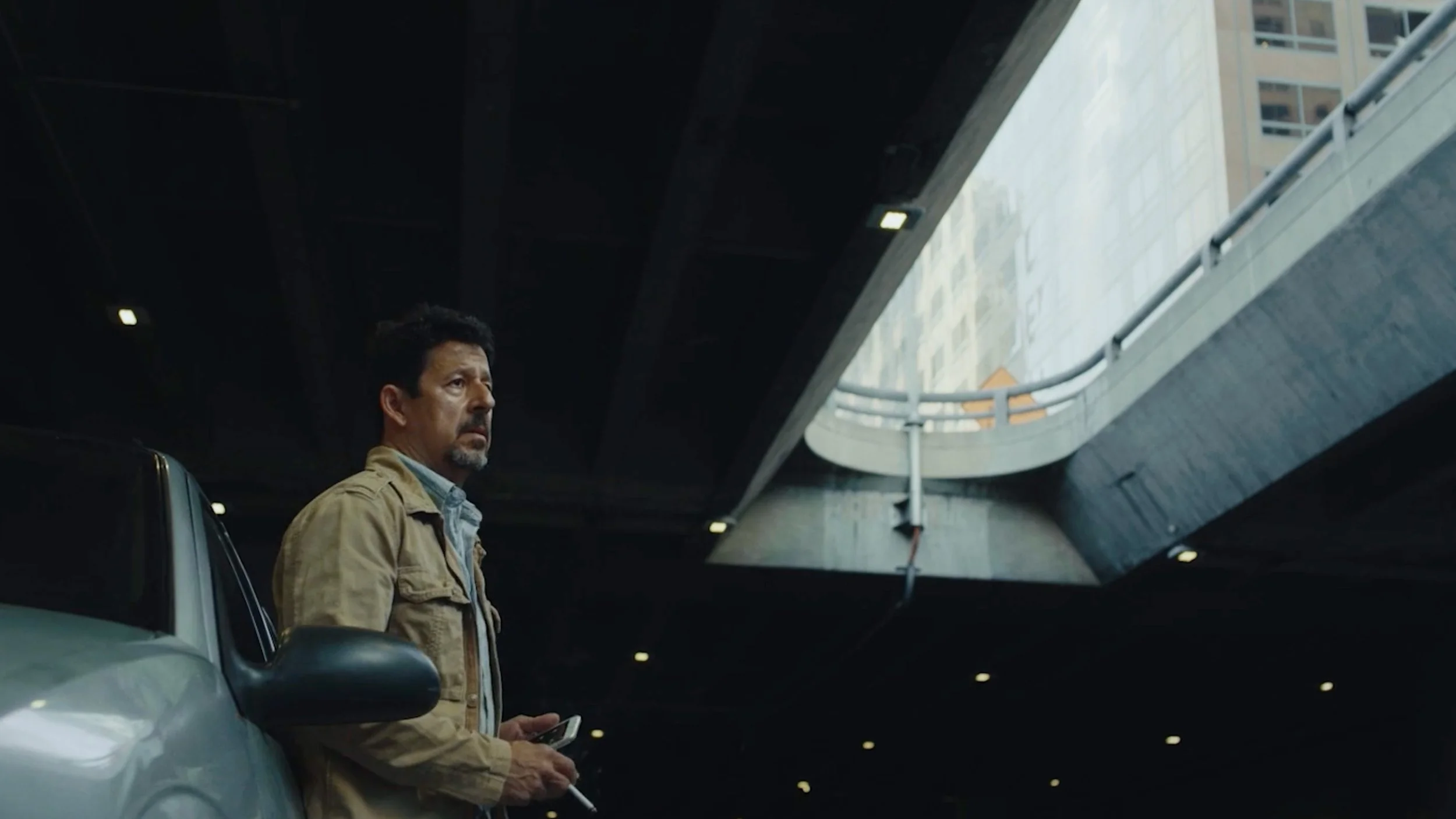 A darkly lit parking garage with a man leaning on a car, holding a cigarette and a phone, looking serious.
