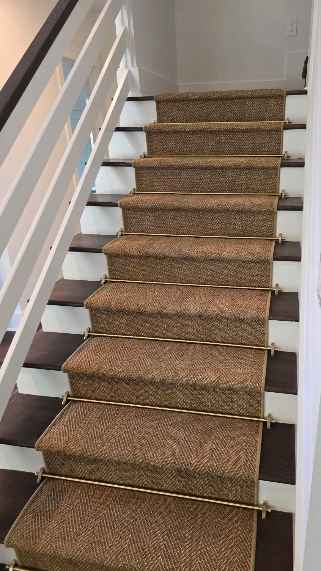 Wooden staircase with a brown carpet runner secured with brass stair rods, white railing on the left, white walls, and a power outlet on the right.