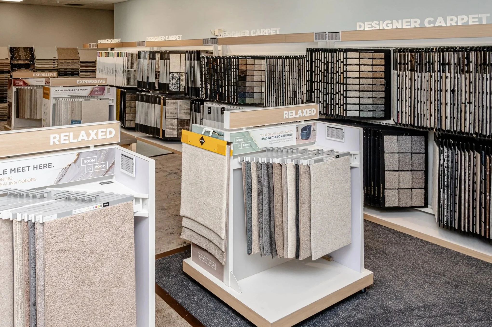 Display of various carpet samples in a flooring store, organized into sections labeled 'Relaxed' and 'Designer Carpet' with a selection of carpet swatches hanging on racks.
