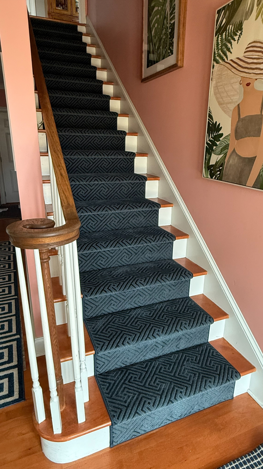 Close-up of a staircase with black patterned carpet, wooden steps, white risers, and a wooden handrail, inside a house with pink walls decorated with framed art.