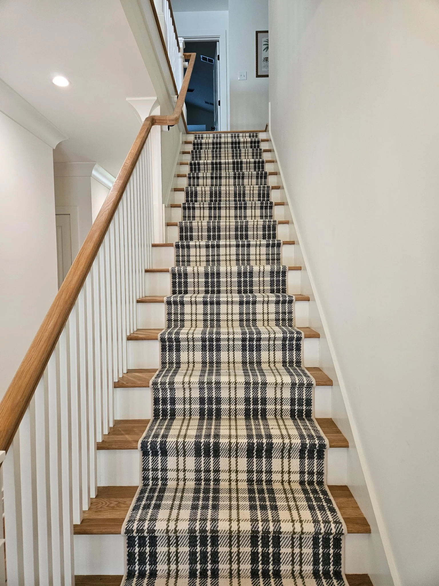 A staircase with a black, white, and gray plaid runner rug, wooden steps, white railings, and walls, leading to an upper floor with a doorway and framed artwork.