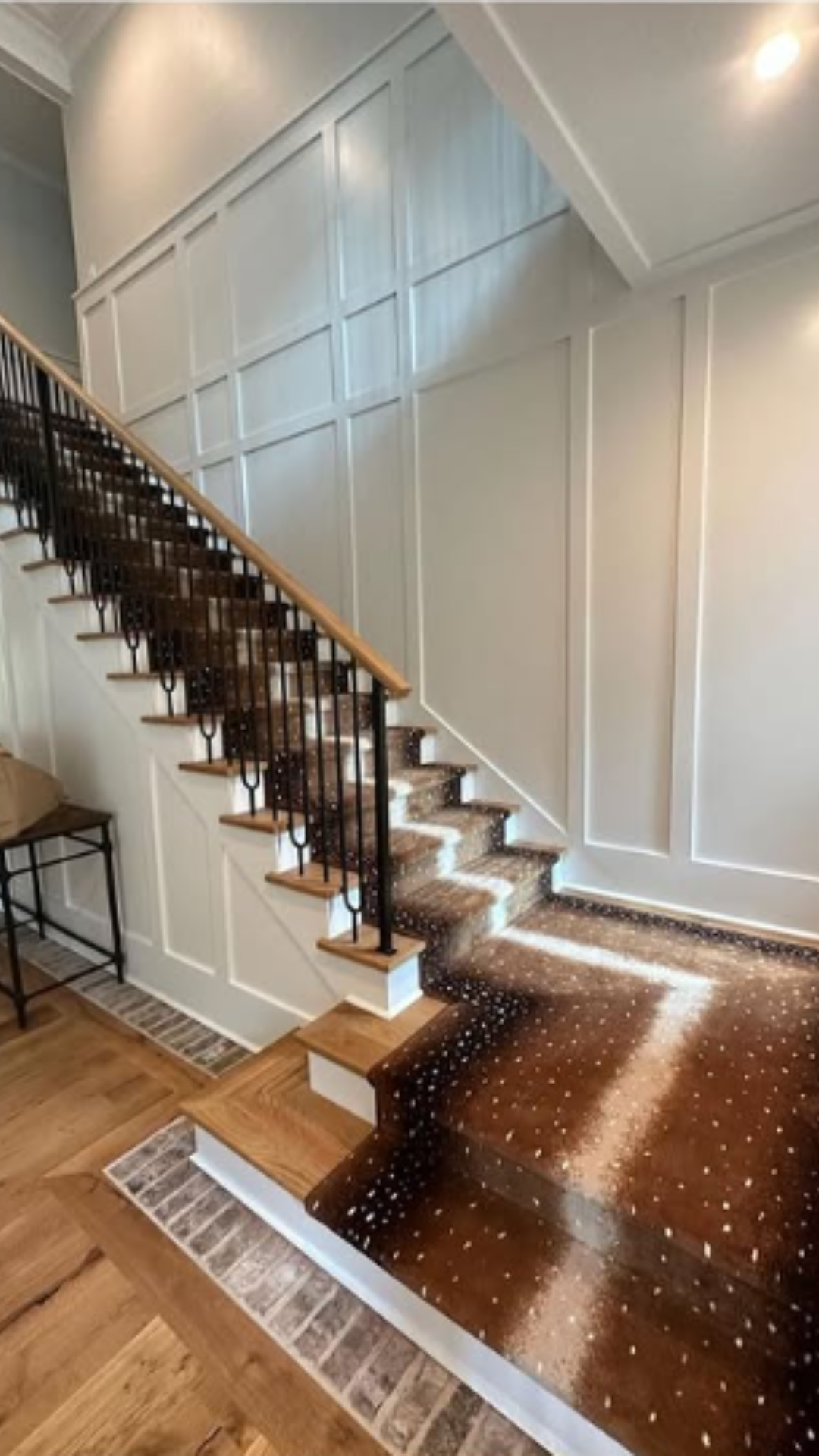 Interior view of a staircase with wooden steps, black metal balusters, and a wooden handrail. The staircase has a patterned carpeted flooring, and the walls are decorated with white paneled wainscoting.