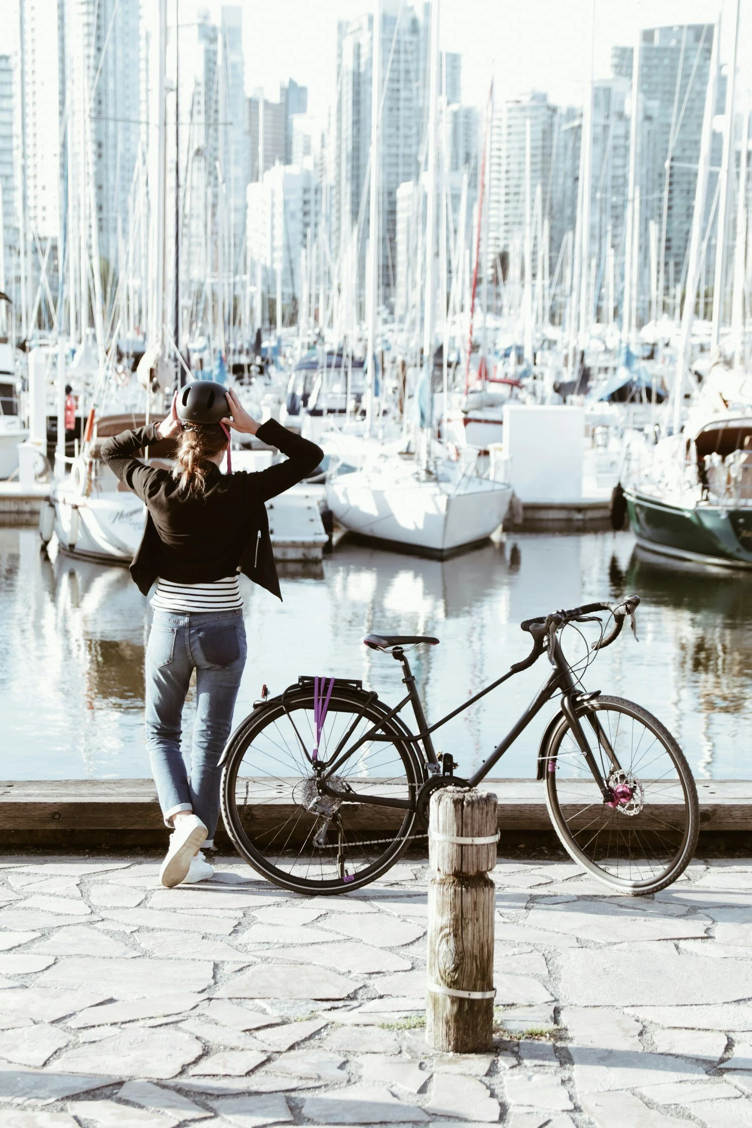 A woman in a black jacket and striped shirt adjusting her helmet near a bicycle, with a marina of sailboats and tall city buildings in the background.