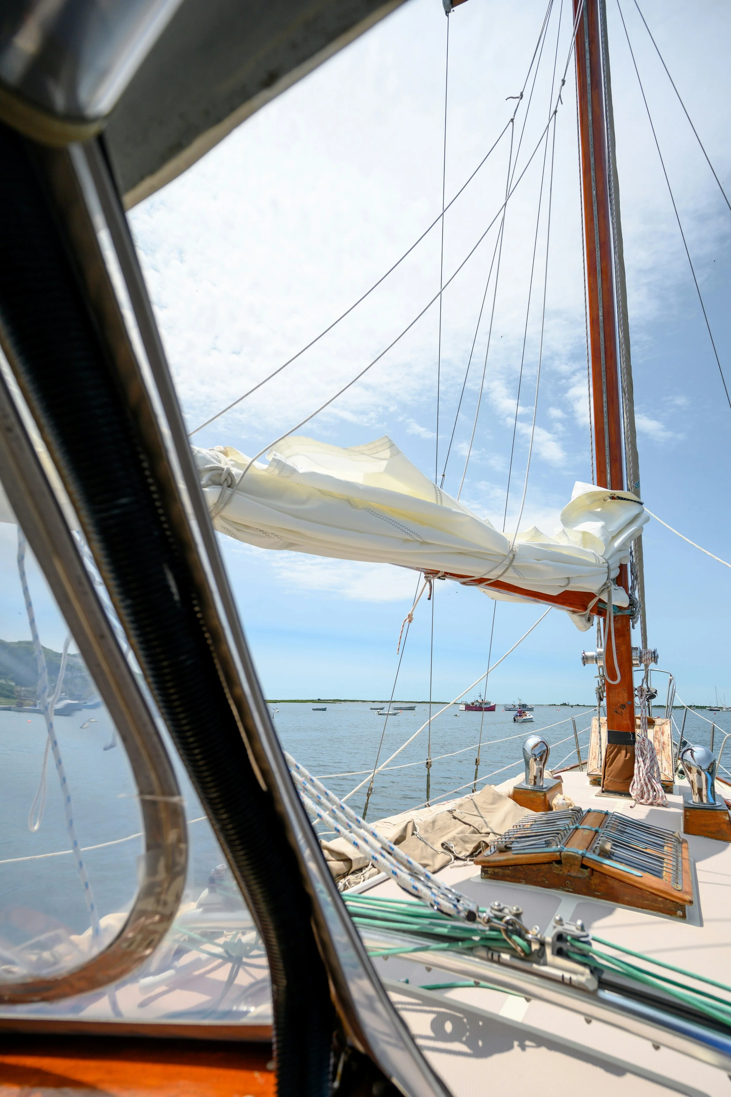 View from a sailboat looking towards the bow with a large white sail up, surrounded by calm water and other boats in the distance under a partly cloudy sky.