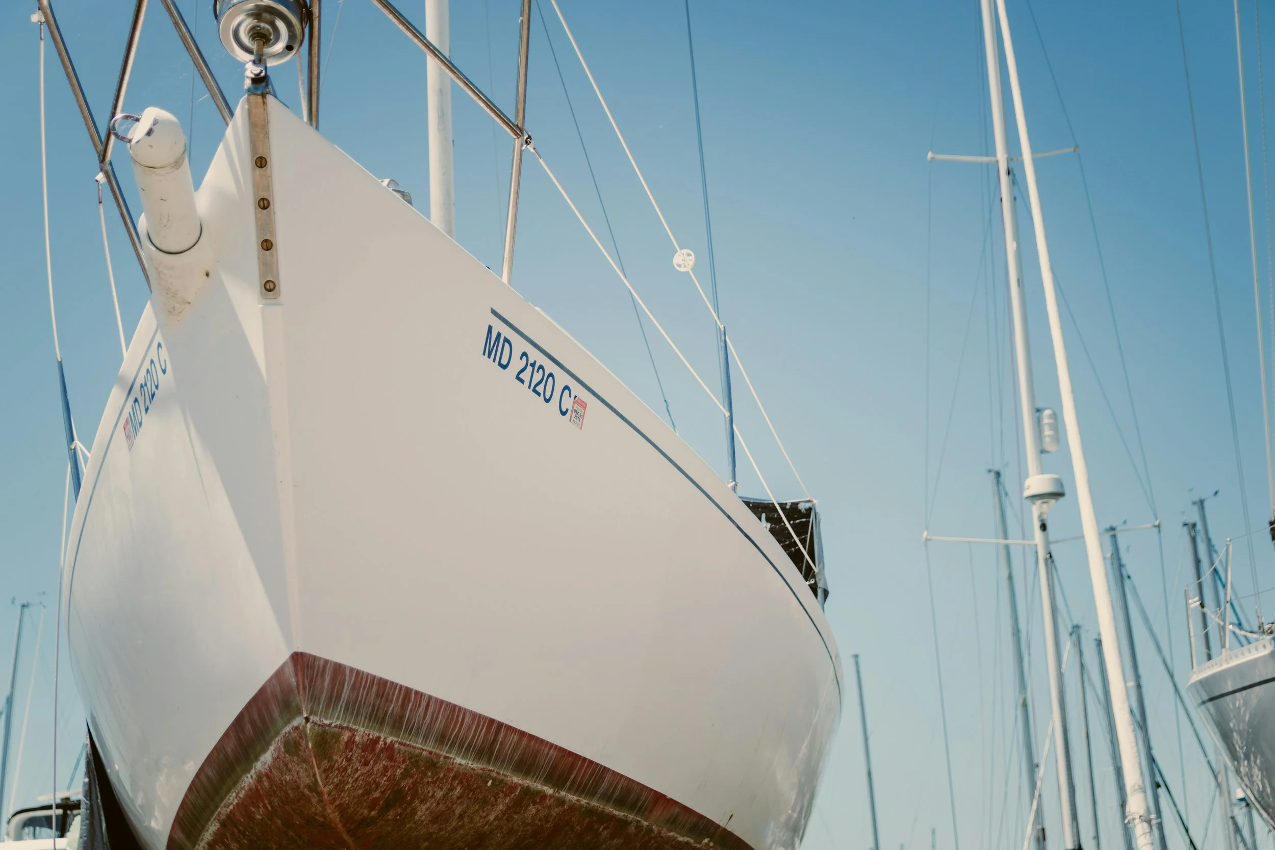 Close-up of the bow of a white sailboat docked at a marina with other sailboats visible in the background under a clear blue sky.