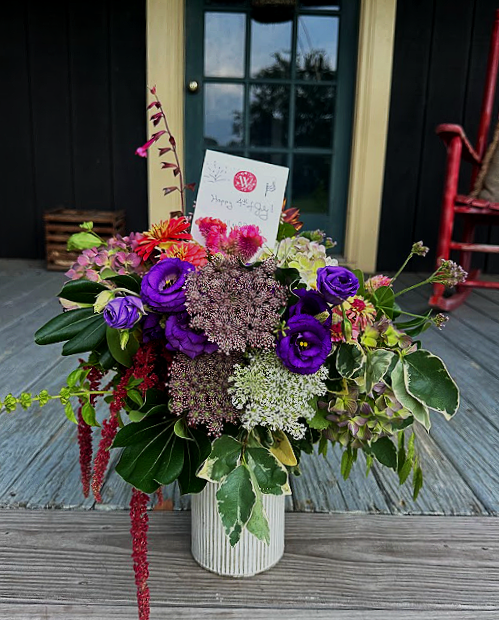 A bouquet of purple, pink, and white flowers in a white vase on a wooden deck, with a black door and window behind it. There is a small card with a heart and a message.