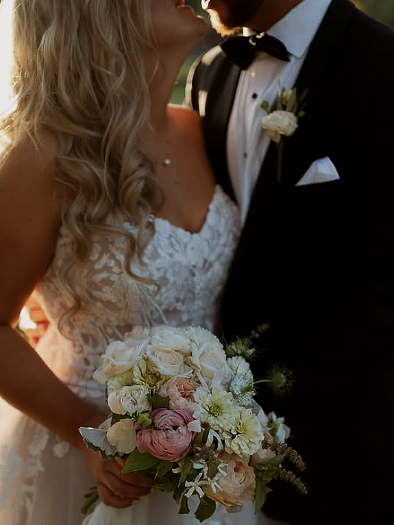 Close-up of a bride and groom on their wedding day, with the bride holding a bouquet of white and pink flowers and the groom in a black tuxedo with a boutonniere.