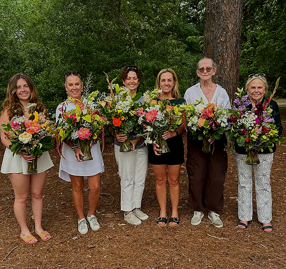 Six women standing outdoors in a wooded area, holding large bouquets of colorful flowers, smiling at the camera.