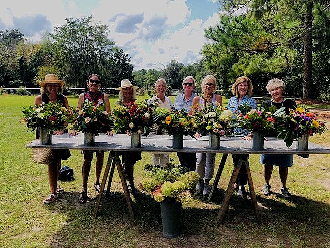 Seven women standing behind a long table with flower arrangements outdoors in a park or garden.