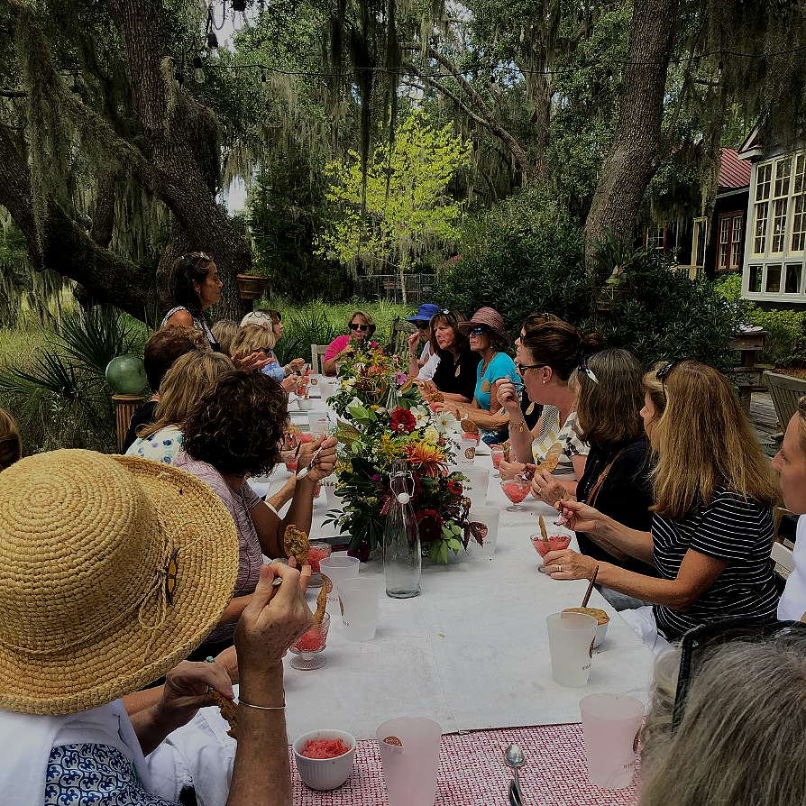 A group of women gathered around a long outdoor table decorated with flowers, enjoying a meal on a sunny day amidst trees and greenery.