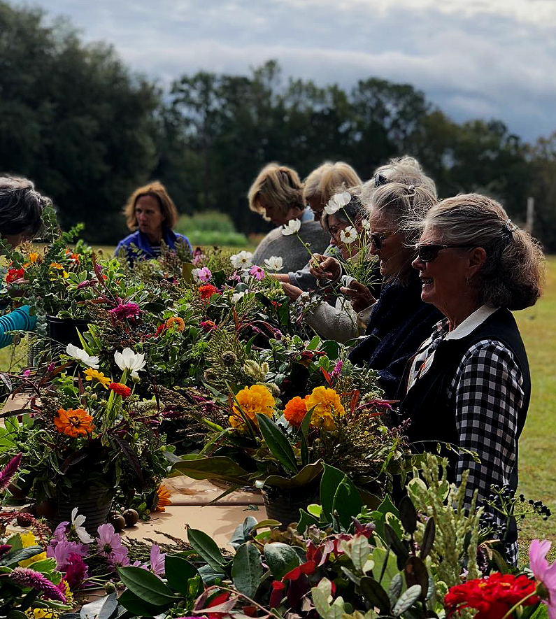 Group of women gathered outdoors, arranging and creating floral arrangements with colorful flowers on a long table of flowers.
