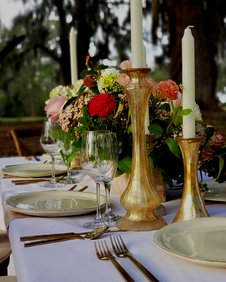 Elegant outdoor dining table with white tablecloth, floral centerpiece, gold candle holders, and empty wine glasses.