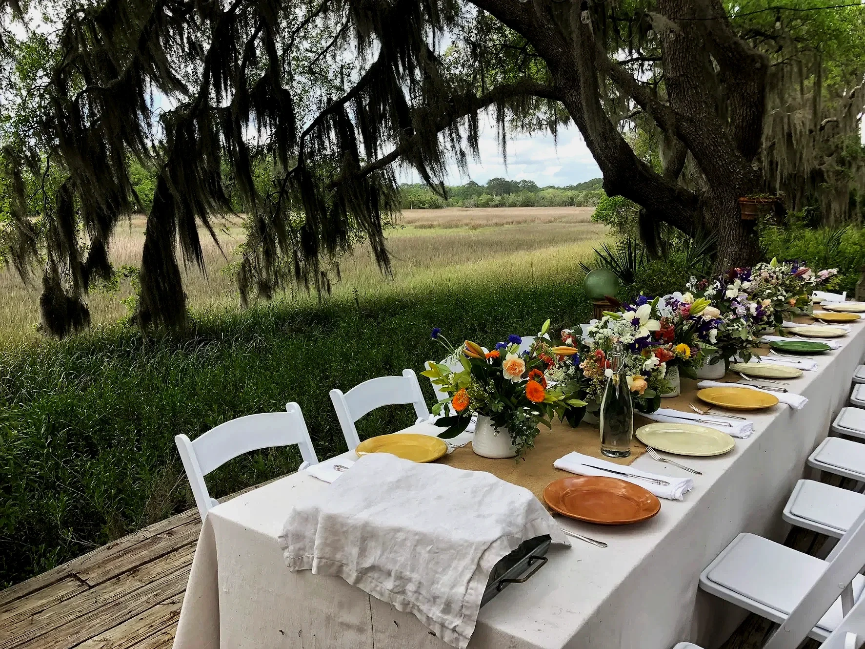 An outdoor dining table set under a large tree with lush greenery and open field in the background, decorated with colorful flower arrangements and tableware.