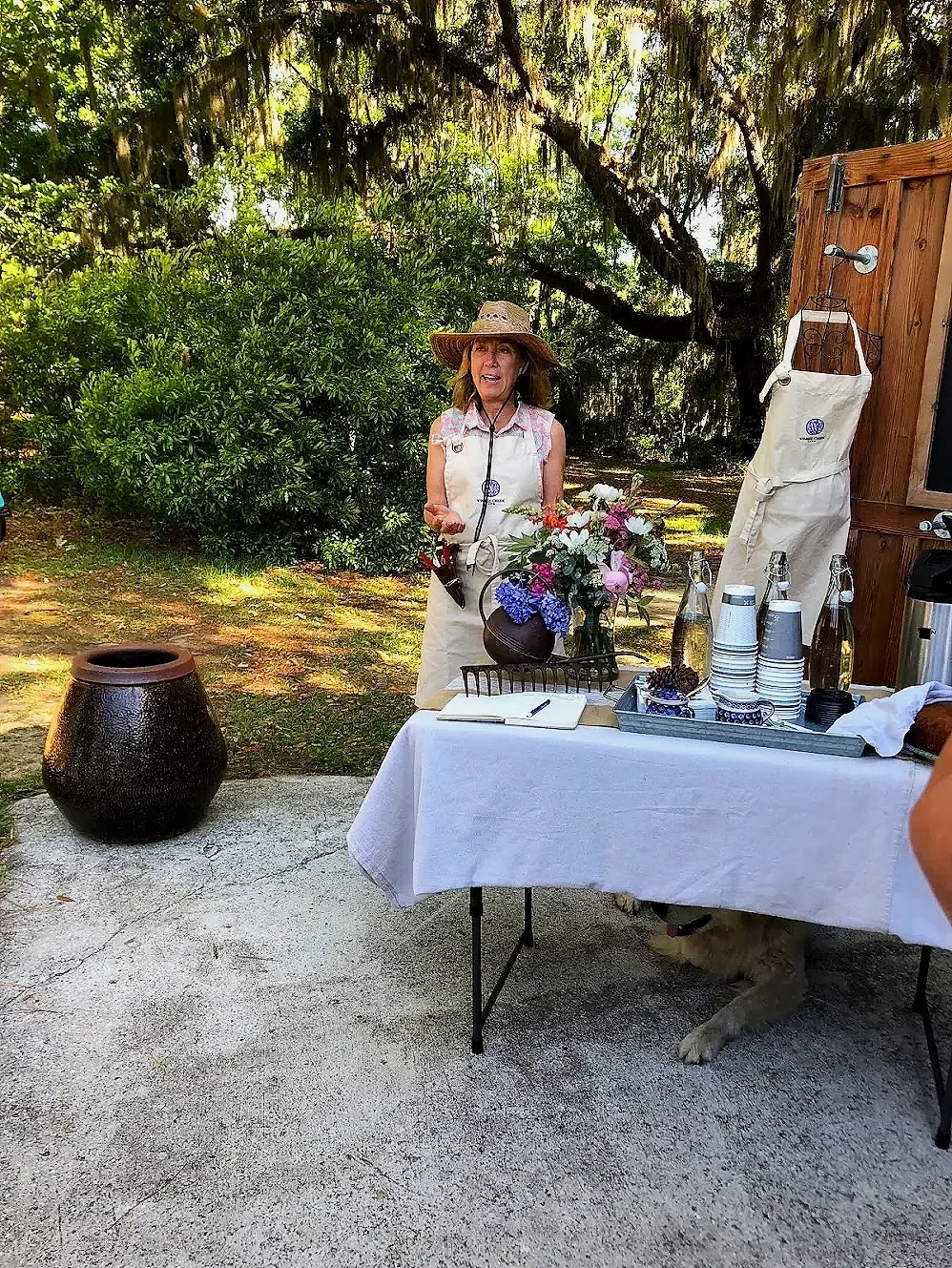 A woman wearing a straw hat and apron stands outdoors near a table covered with a white cloth, decorated with a vase of flowers. She appears to be speaking or explaining something. There are cups, water bottles, and a notepad on the table, and trees 