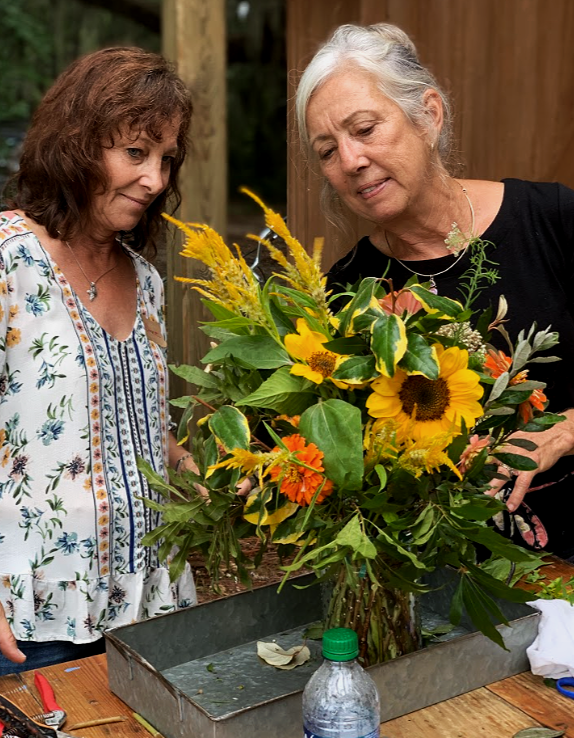 Two women are arranging a large bouquet of sunflowers, orange flowers, and greenery in a metal container on a wooden table outdoors.