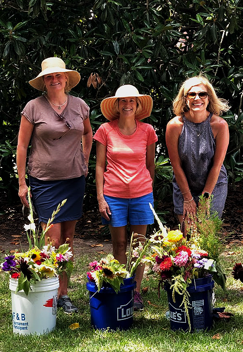 Three women standing outdoors behind three buckets filled with colorful flowers, wearing summer hats and casual clothing, smiling.