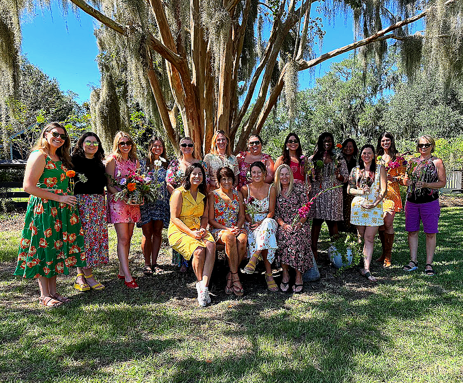 A group of women gathered outdoors under a large tree, some seated and others standing, celebrating with flowers and smiling.