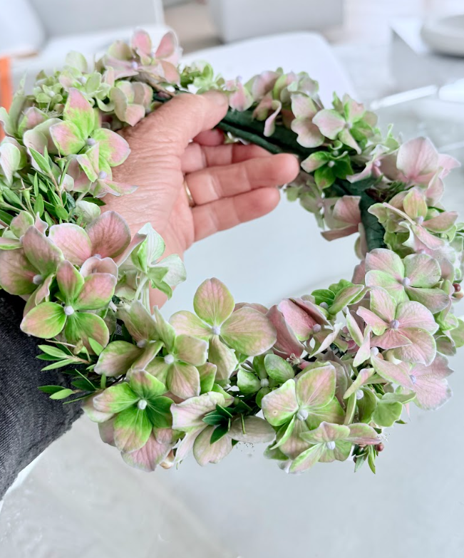A hand holding a pink and green floral wreath made of hydrangea flowers.