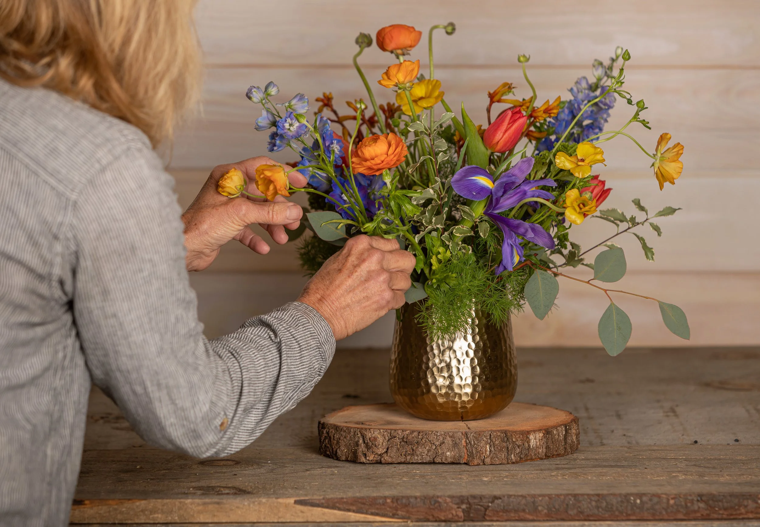A person arranging a colorful flower bouquet in a brown hammered metal vase on a wooden table.
