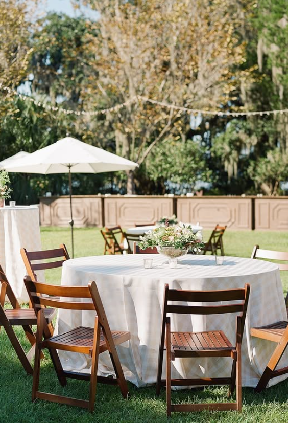 Outdoor event setup with round table, white tablecloth, flower centerpiece, wooden chairs, large umbrella, and a wooden fence in background.