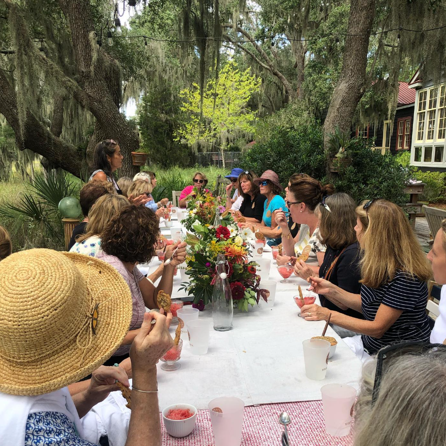A group of women seated at a long outdoor table, having a meal in a lush garden setting with trees and greenery around.
