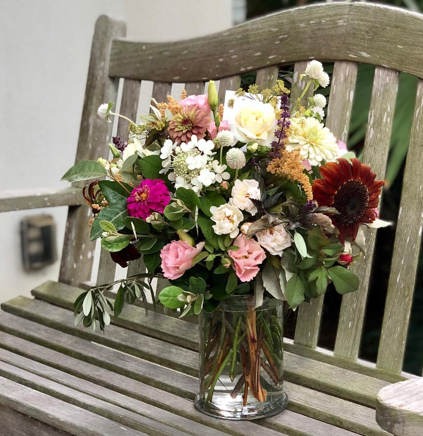 A colorful bouquet of mixed flowers in a glass vase placed on a weathered wooden bench outdoors.