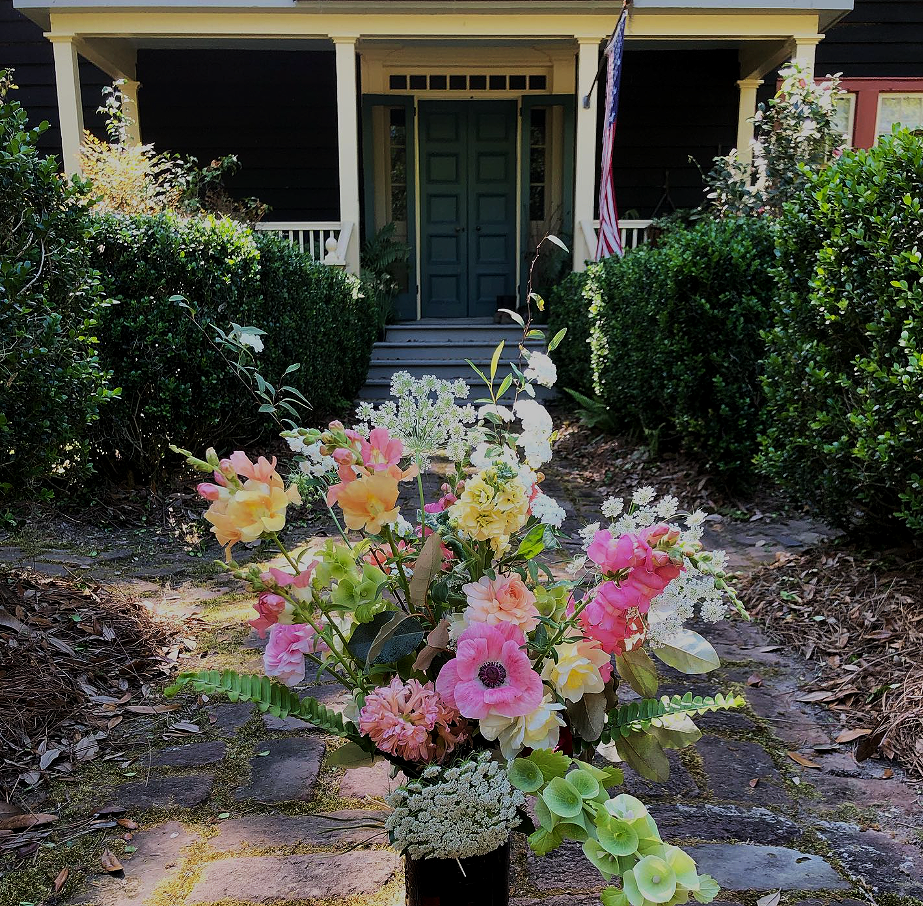 A bouquet of pink, yellow, and white flowers in front of a house with dark green door, steps, and a porch decorated with an American flag.