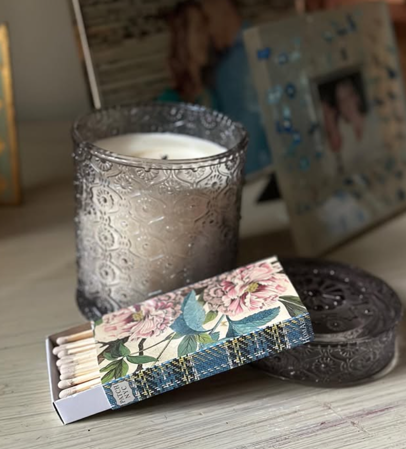 A lit white candle inside a decorative, textured glass holder, a box of matches with floral design, and a decorative dish on a light wooden surface, with a blurred photo frame in the background.