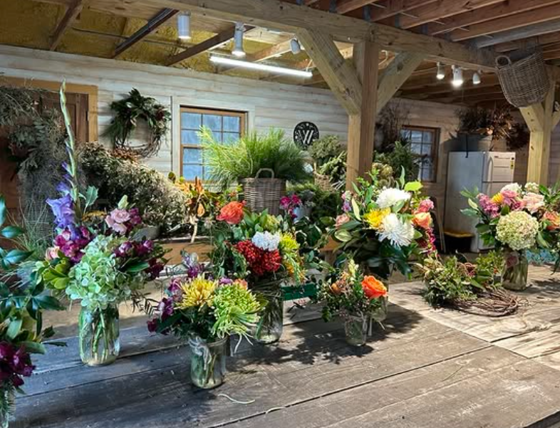 Various colorful flower arrangements in vases on a rustic wooden table inside a barn or workshop.