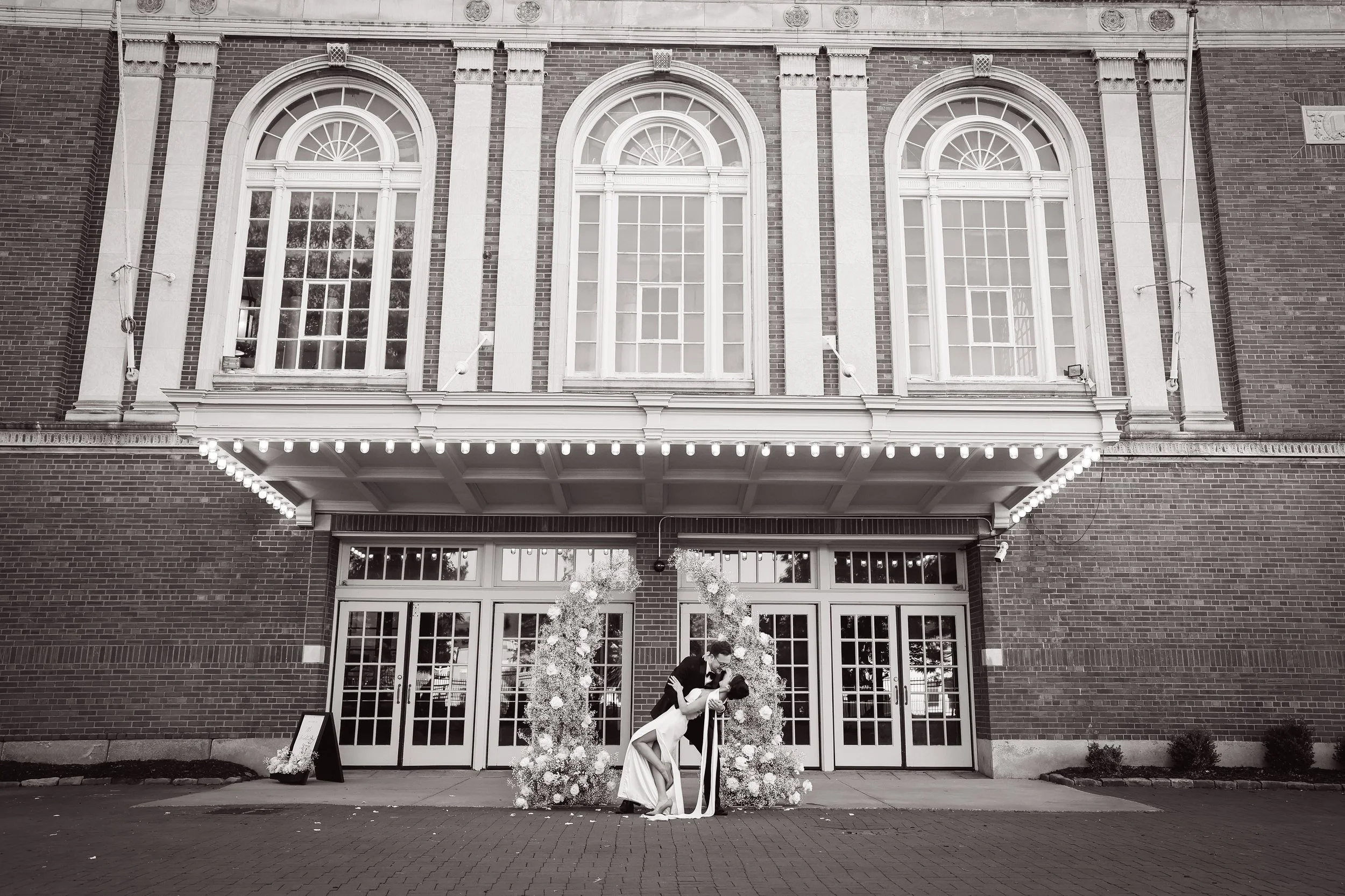 A couple embracing and kissing in front of a decorated floral arch outside a brick building with large arched windows at night.