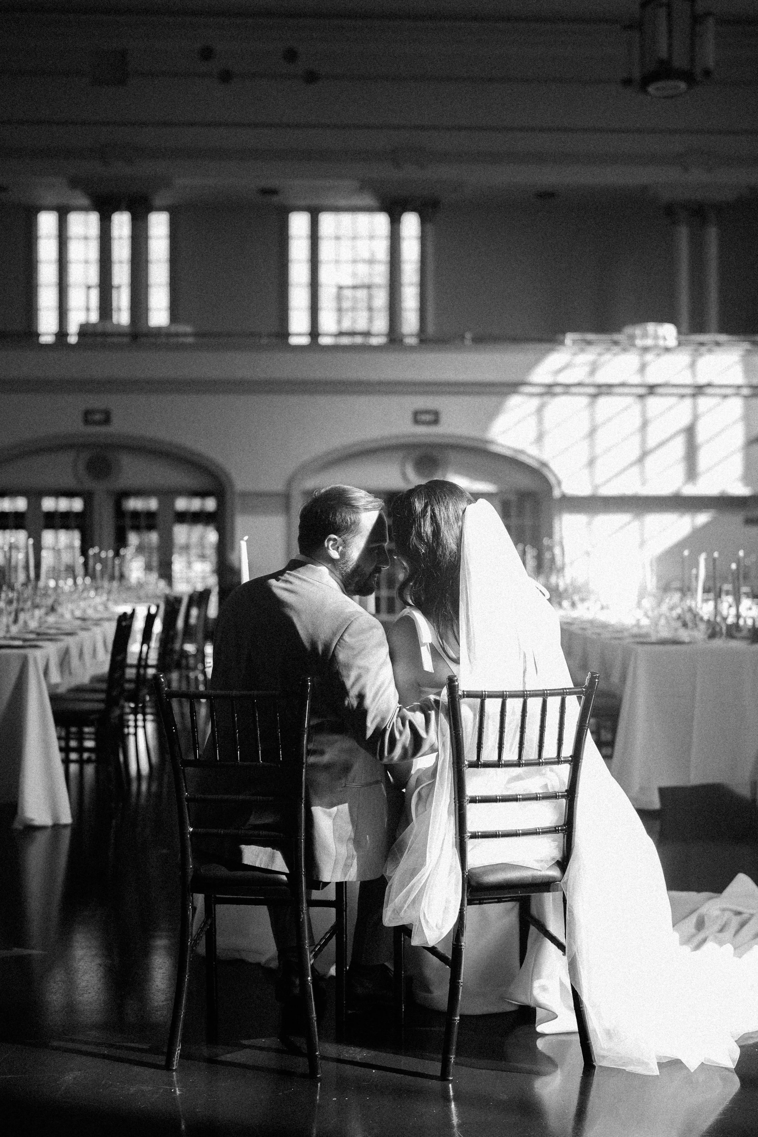 A bride and groom sitting closely together at a wedding reception, sharing an intimate moment. The venue is decorated with long tables, and the lighting creates a warm, inviting atmosphere with sunlight streaming through large windows.