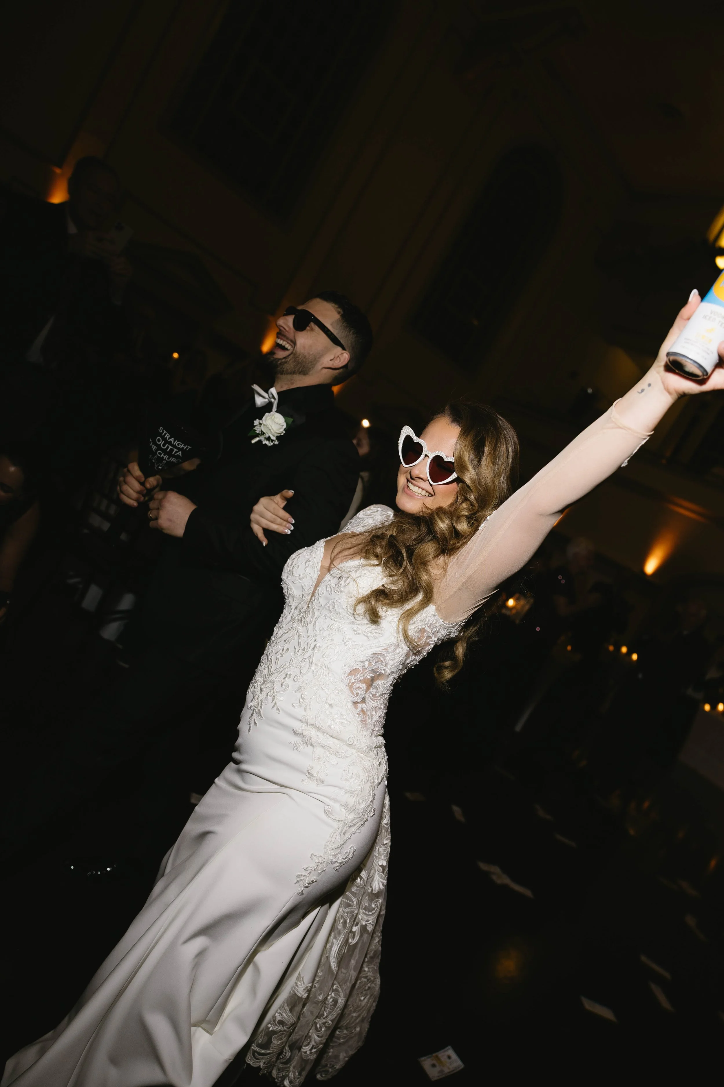 Bride in a white lace dress with heart-shaped sunglasses celebrating at a wedding reception, holding a drink high, dancing and smiling, with a groom in a dark suit in the background.