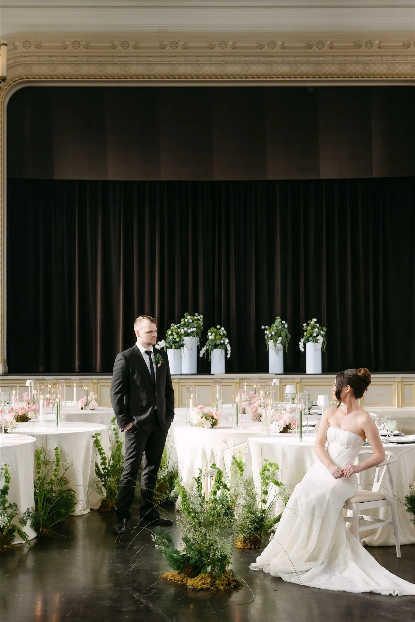 A bride and groom in a wedding ceremony in a large hall with decorated tables and floral arrangements, the groom in a black suit standing and the bride sitting in a white wedding dress.