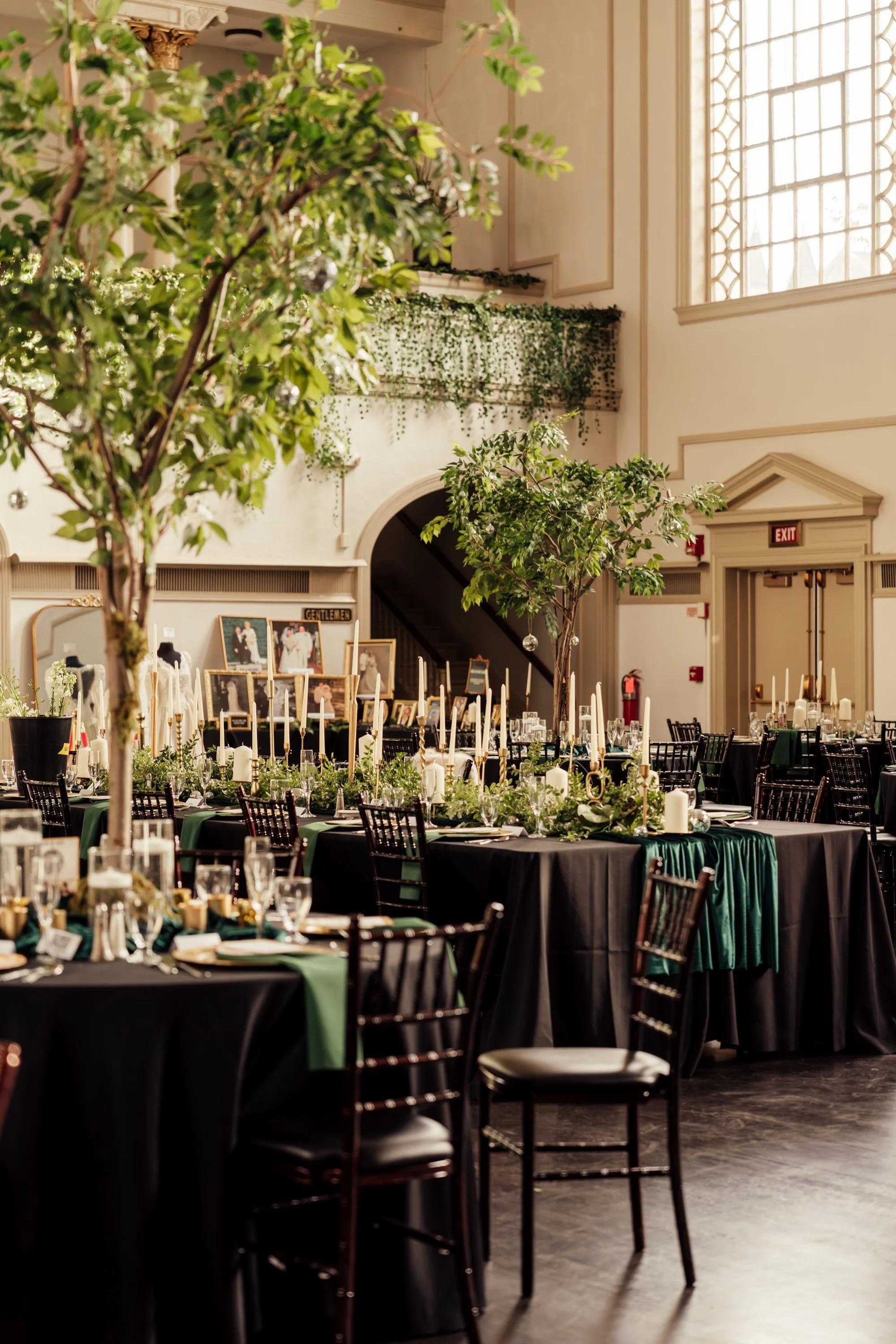 Elegant banquet hall decorated for a formal event with black-clothed tables, greenery, candles, and tall trees, in a grand room with high ceilings and large windows.