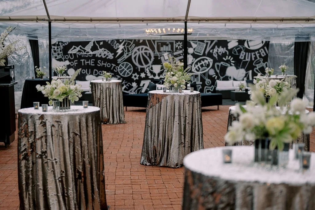 Indoor event space decorated with tall tables covered in silver, tree bark-patterned tablecloths, floral centerpieces, and a black mural with sports-themed drawings and words in the background.