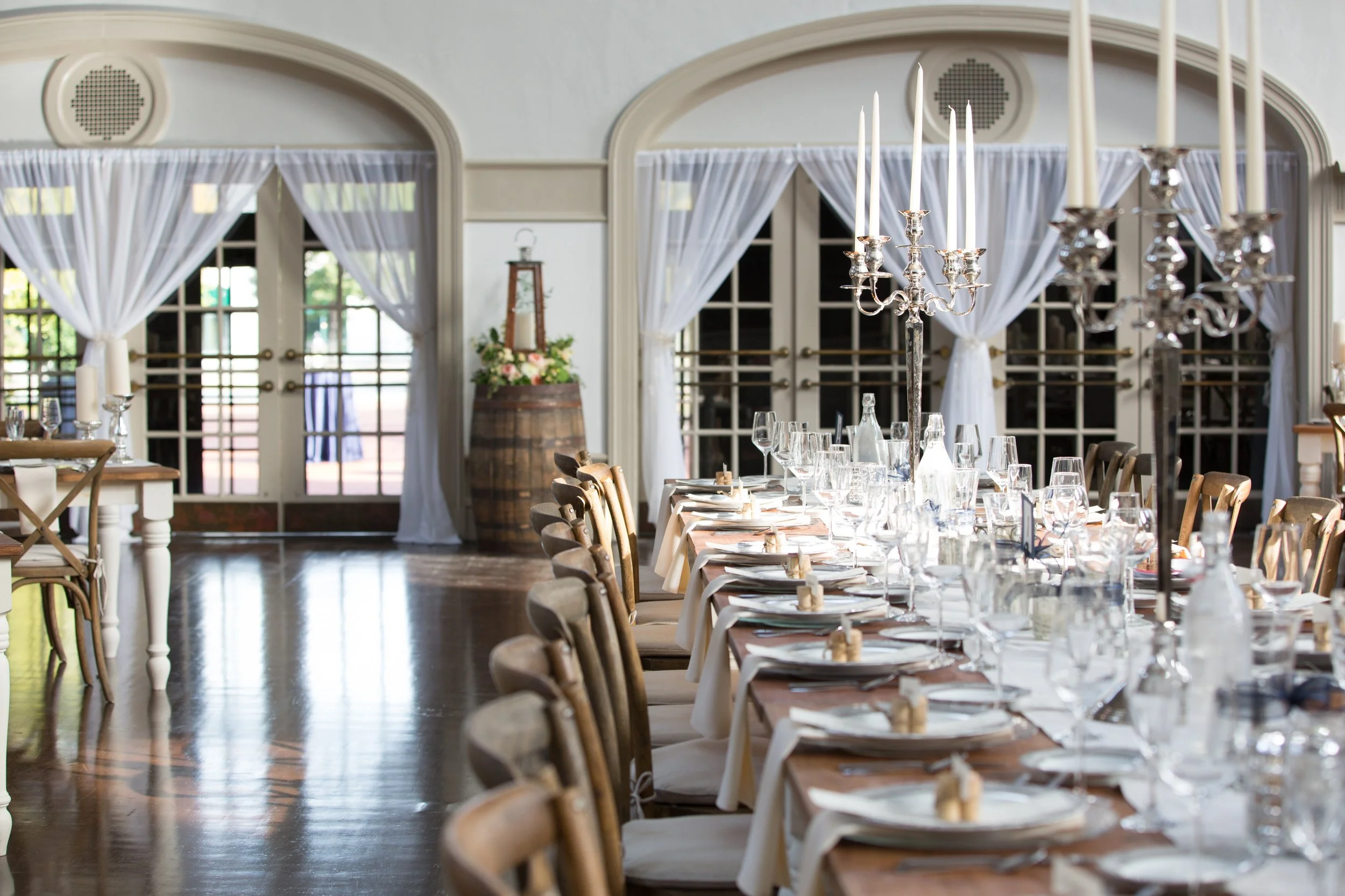 Elegant banquet table set with wine glasses, plates, and candles in a bright, decorated room with large windows and white curtains.