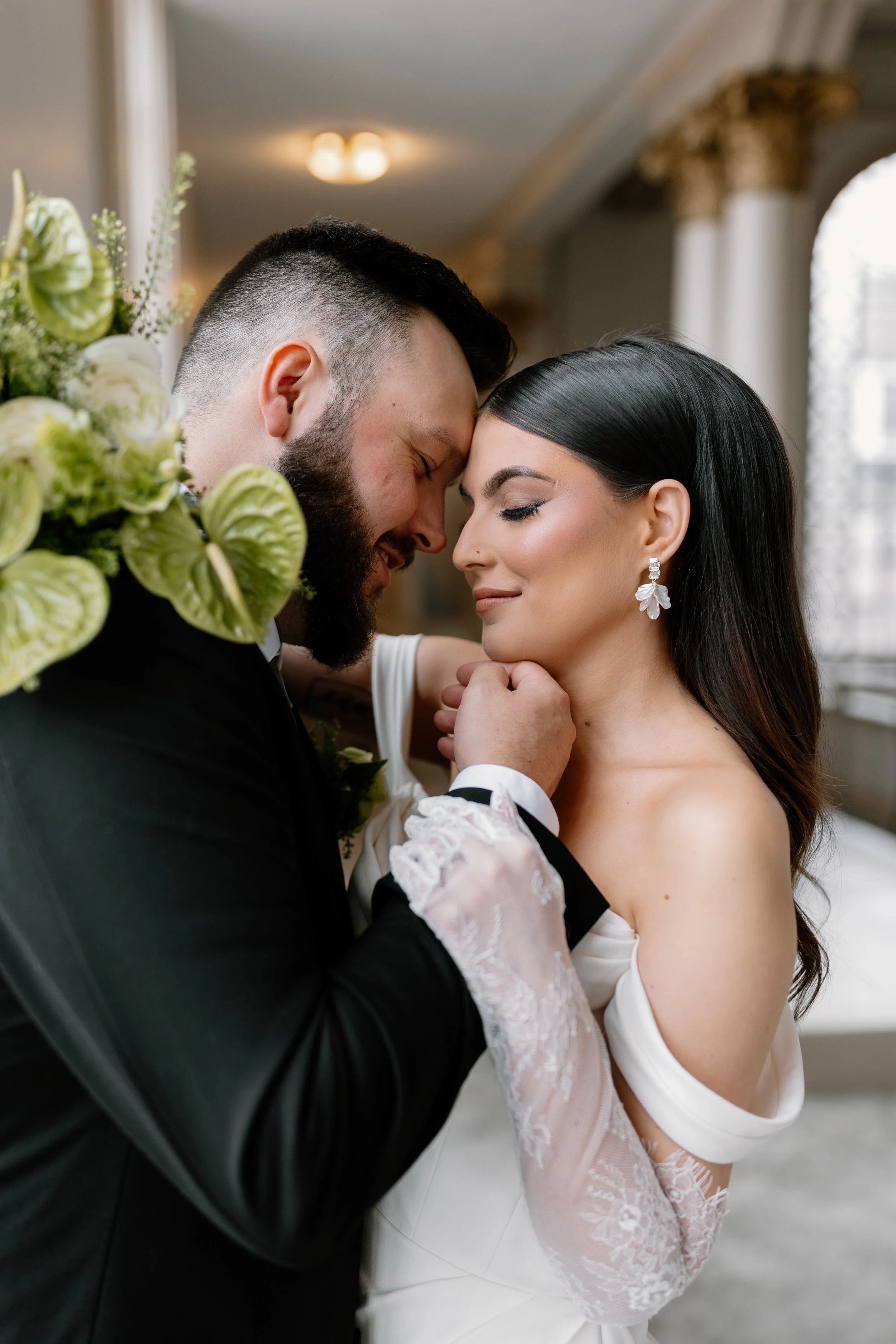 Close-up of a newlywed couple embracing with foreheads touching, eyes closed, in a decorated room.