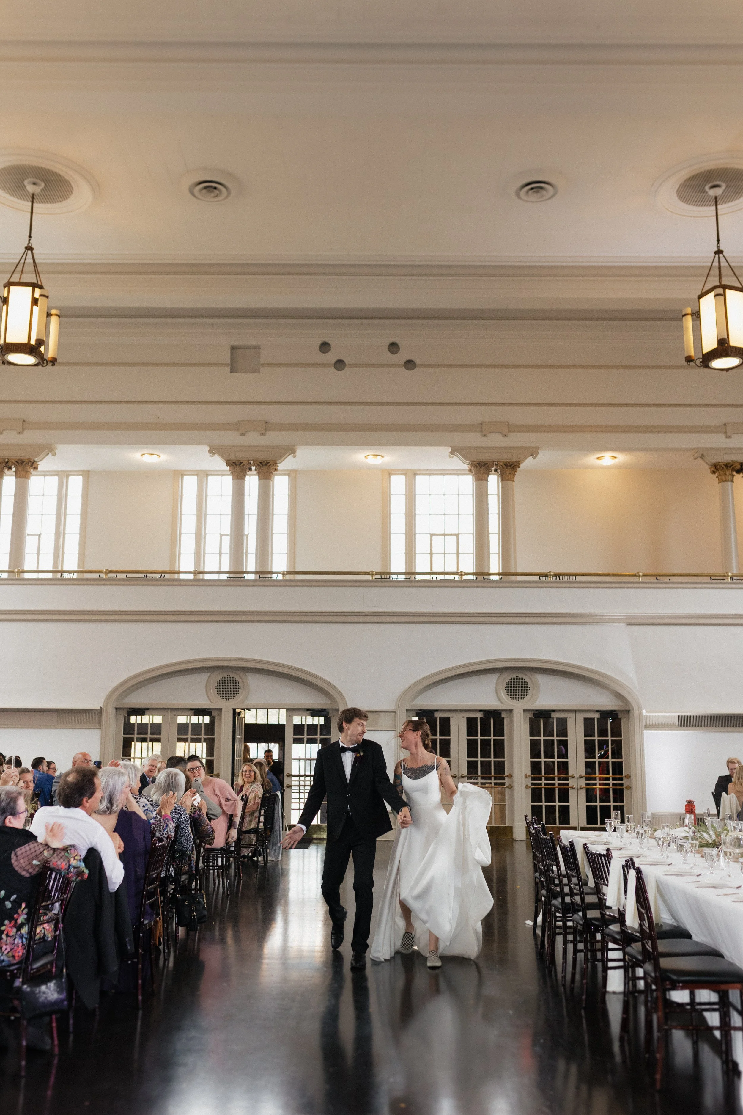 A bride and groom walking hand-in-hand down the aisle of a wedding reception hall, with guests seated on either side at decorated tables, celebrating their wedding.