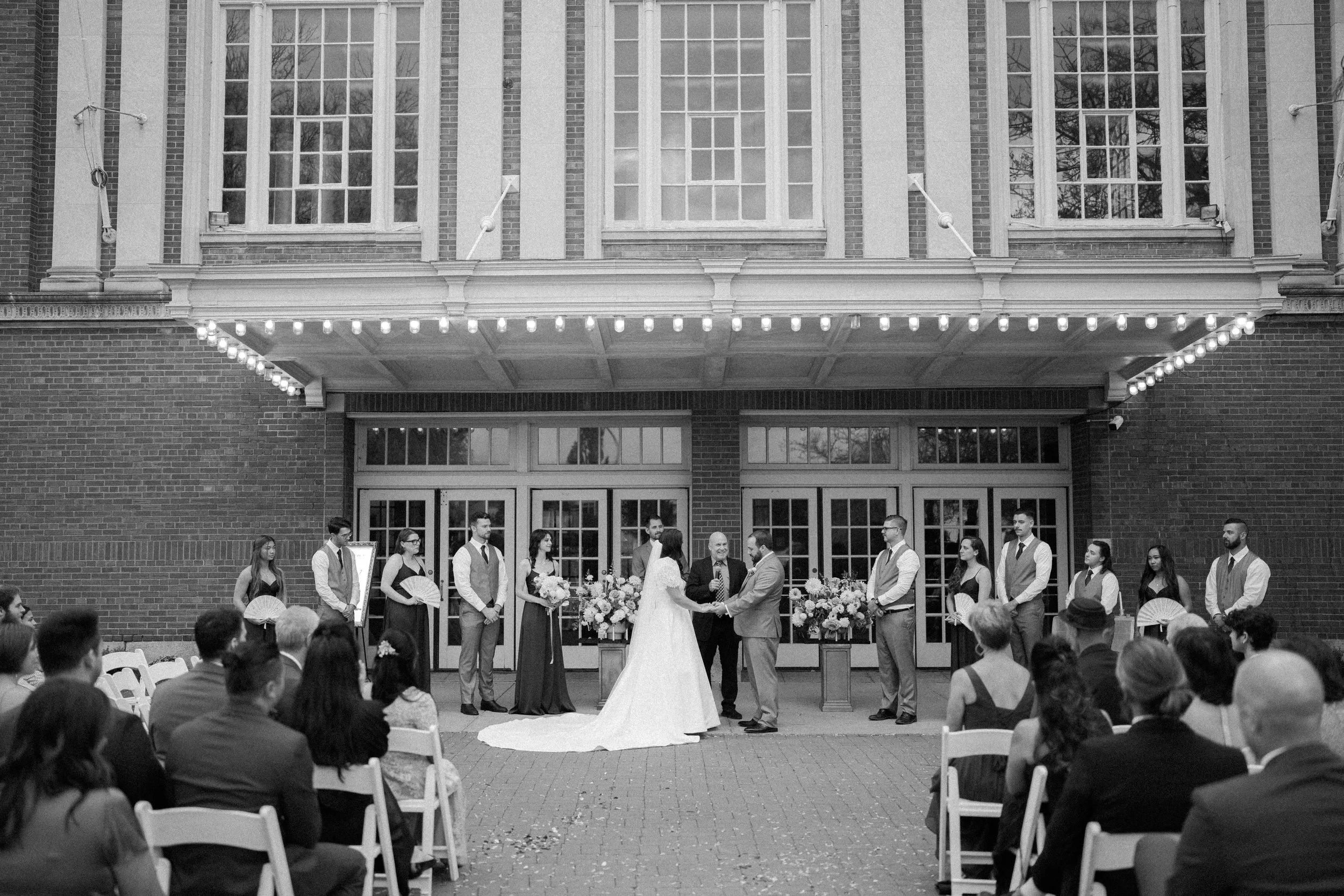 A wedding ceremony taking place outdoors in front of a brick building with large windows. The bride and groom are holding hands and facing each other, surrounded by attendants and seated guests. The bride is dressed in a white gown and veil, and the groom in a suit. Flowers and decorations are arranged around the ceremony area.