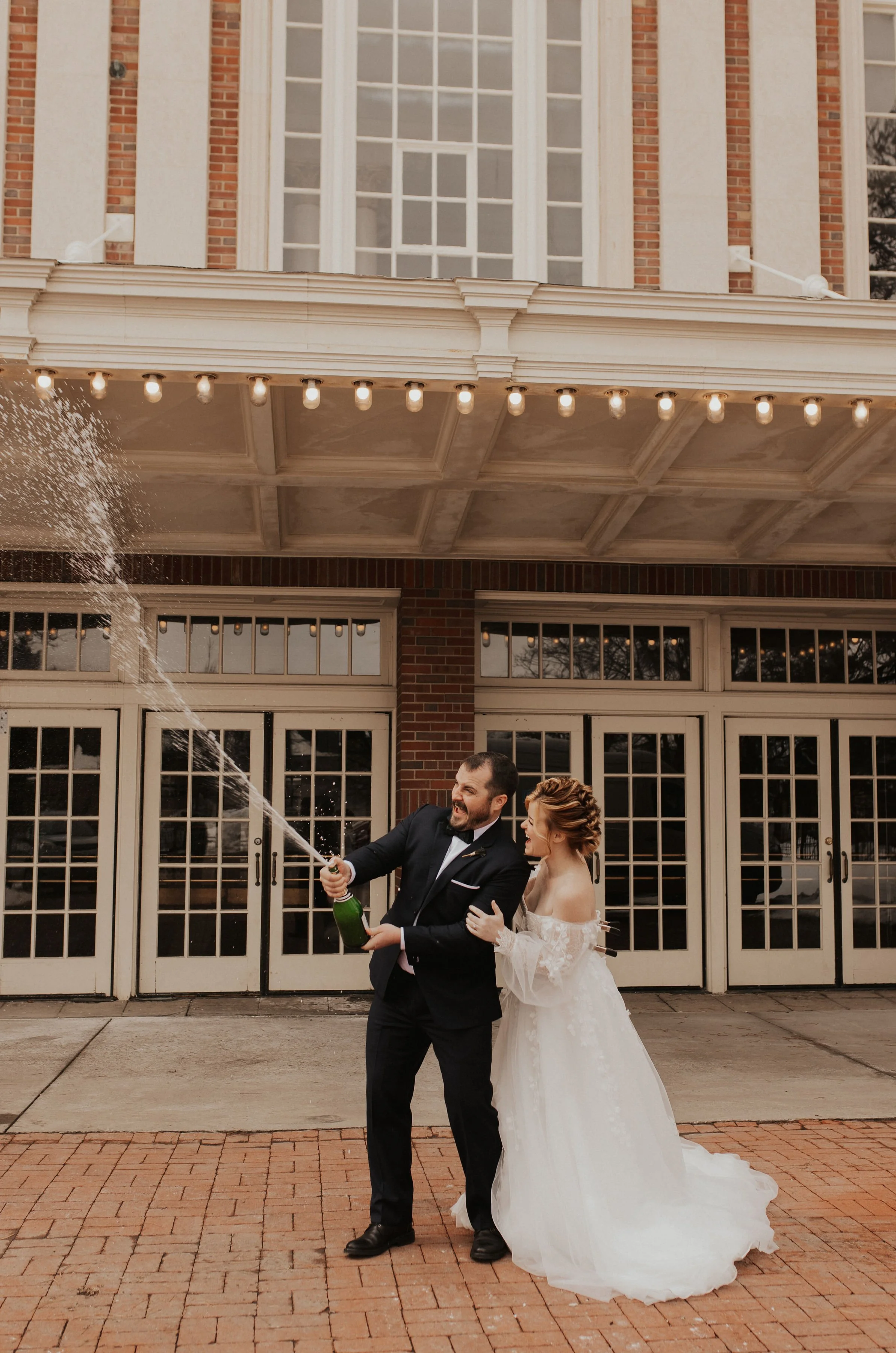 A newlywed couple celebrating outside a building, with the groom opening a champagne bottle and spraying it, while the bride smiles and holds onto him.