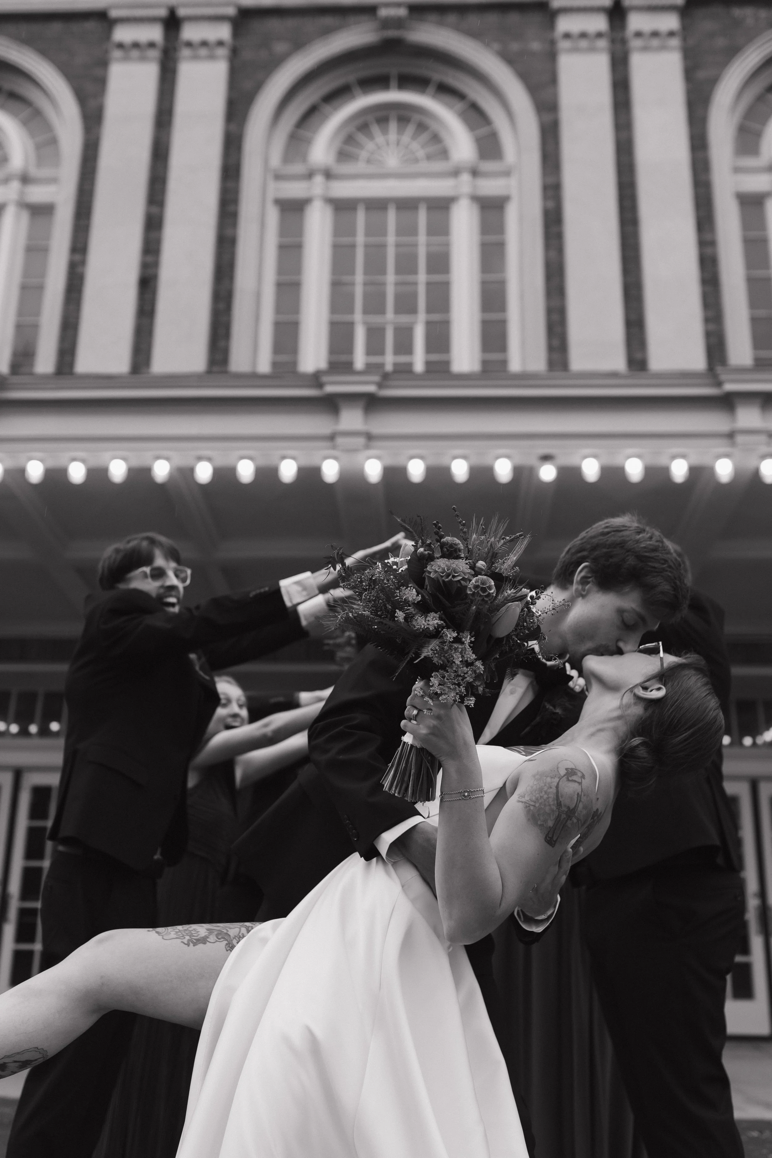 A couple sharing a kiss at their wedding, with the bride holding a bouquet of flowers and leaning back, while the groom dips her. Several friends in tuxedos are spraying champagne behind them. The scene takes place in front of a grand, illuminated building with tall windows.