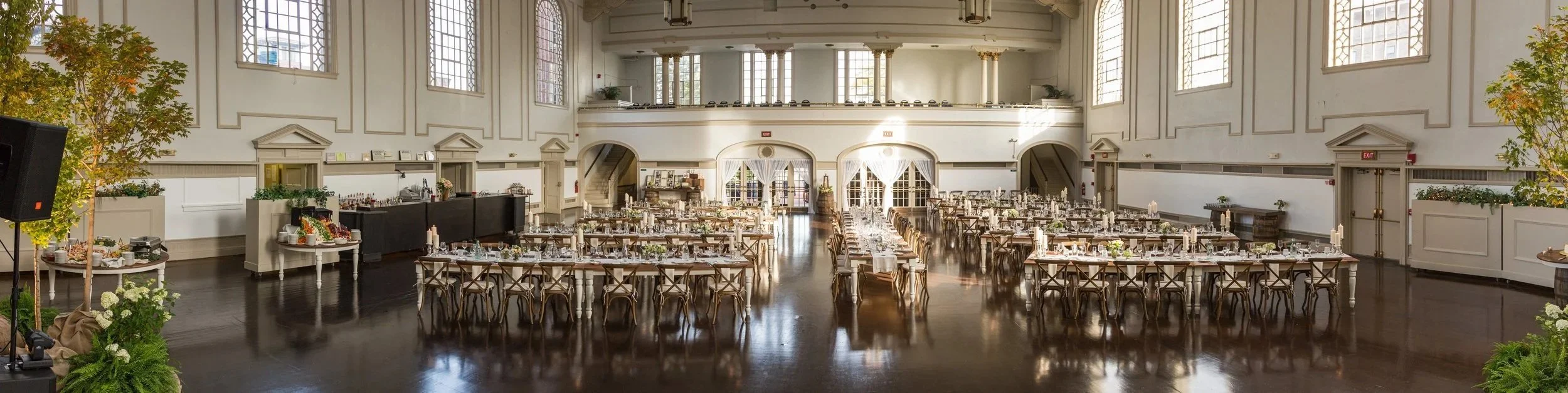 Elegantly decorated banquet hall set up for a wedding reception with long tables, chairs, candles, and floral centerpieces, featuring high ceilings and large windows.