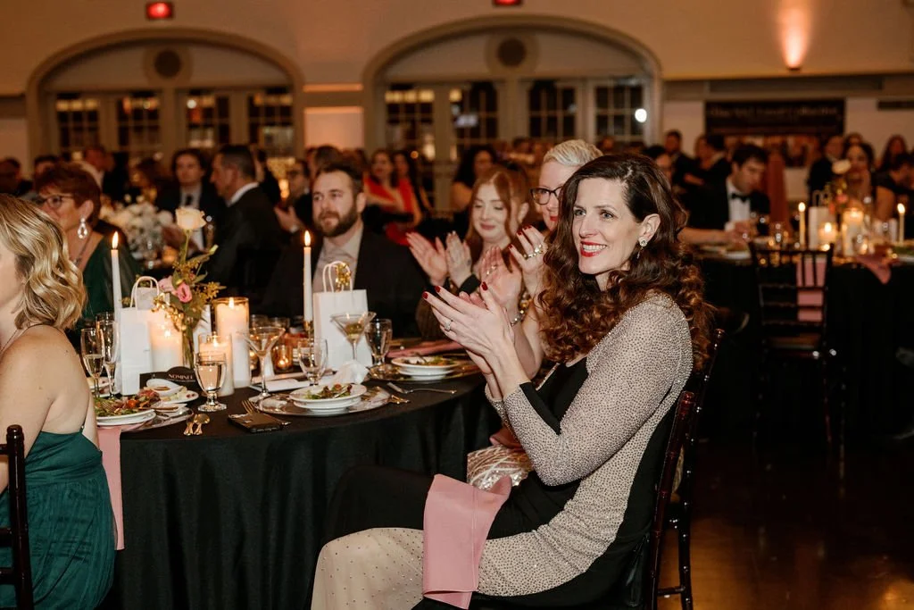 People sitting at a banquet table with lit candles, flowers, and elegant dinnerware, clapping and smiling during a formal event in a decorated hall.