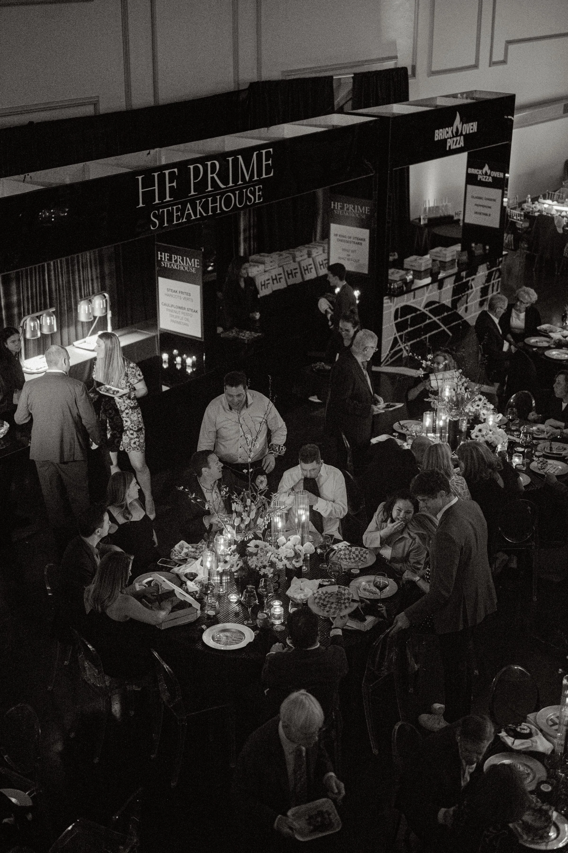 People dining at a formal event with tables decorated with flowers and candles, and food boxes on the tables. In the background, there is a stand with signs for HF Prime Steakhouse and Brick Oven Pizza.