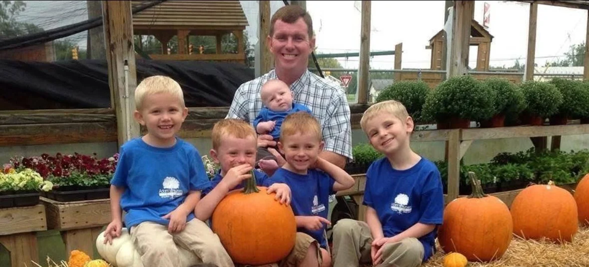 Group of young boys and a man at a pumpkin patch, holding pumpkins and smiling.